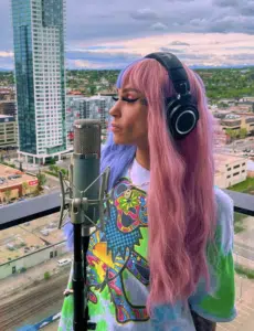 Person with long pastel pink and purple hair, wearing headphones and a colorful shirt, stands in front of a microphone on a city balcony with buildings and cloudy sky in the background.